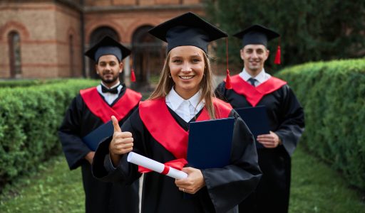 Graduate female shows like with her friends in graduation gowns holding diploma and smiling at camera.