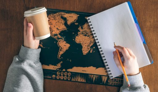 A paper cup in female hands, an empty notepad and a world map on a wooden table. Travel and education concept.